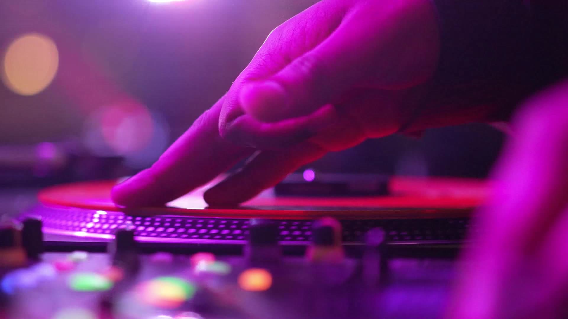 A DJ's hand cueing a spinning vinyl record under pink and purple club lights